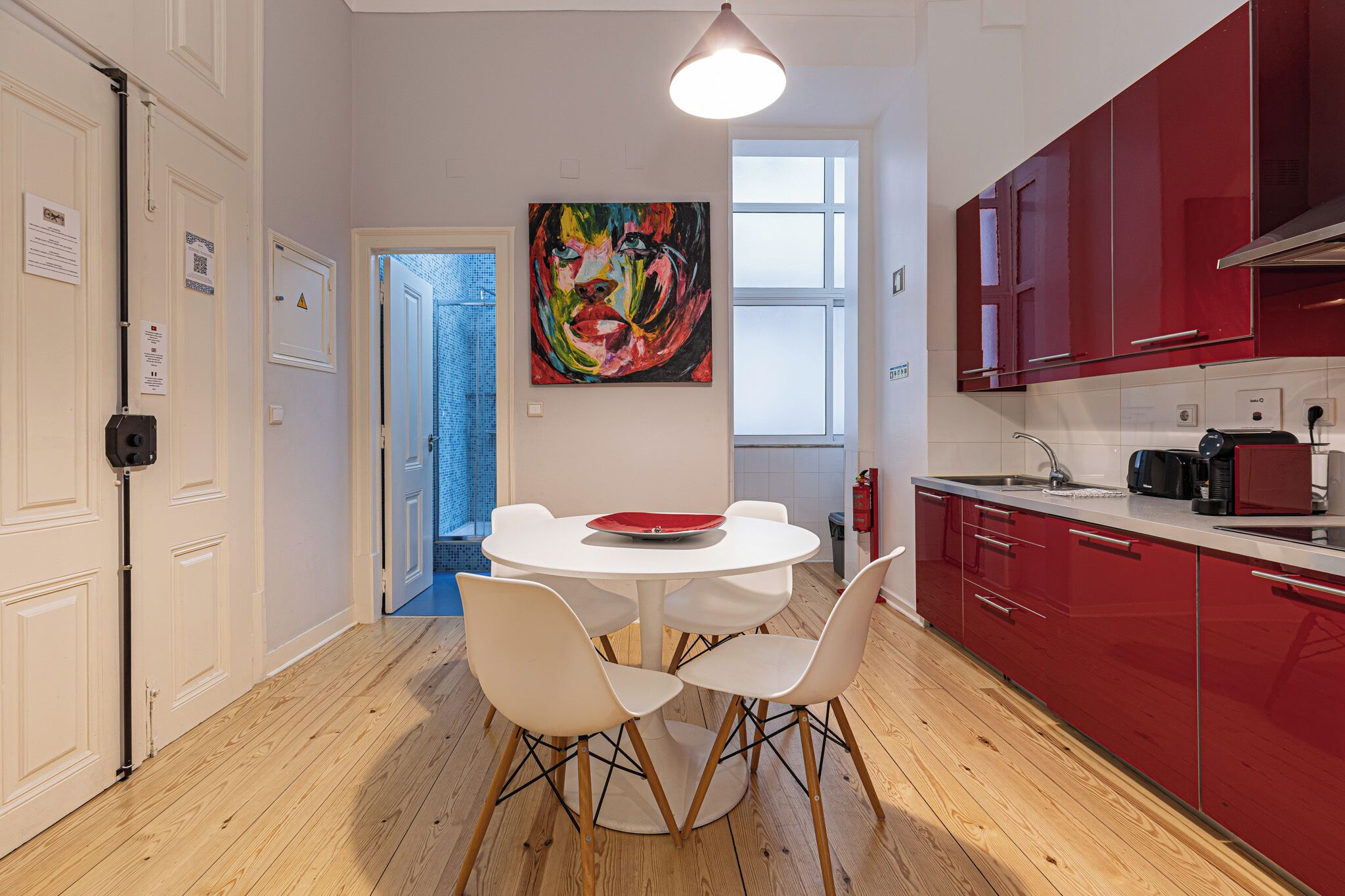 A modern kitchen with red cabinets, a white dining table with four chairs, and a colorful abstract painting on the wall.