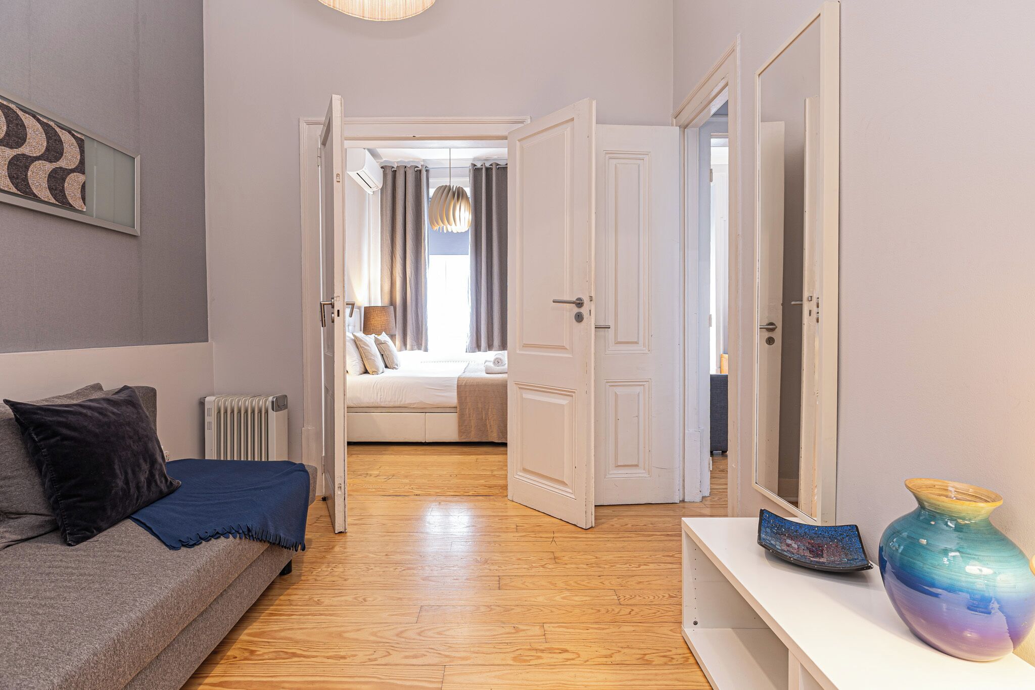 A modern room with a grey sofa, a blue throw blanket, a white radiator, and a large vase on a white console table.