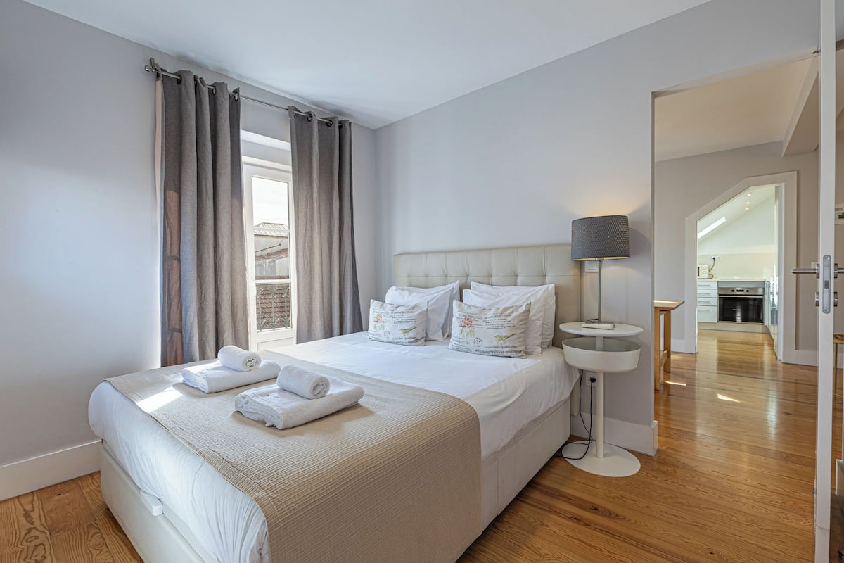 Quarto de hotel com uma cama grande, uma mesa de cabeceira com luminária e vista para a cozinha através de uma porta aberta.