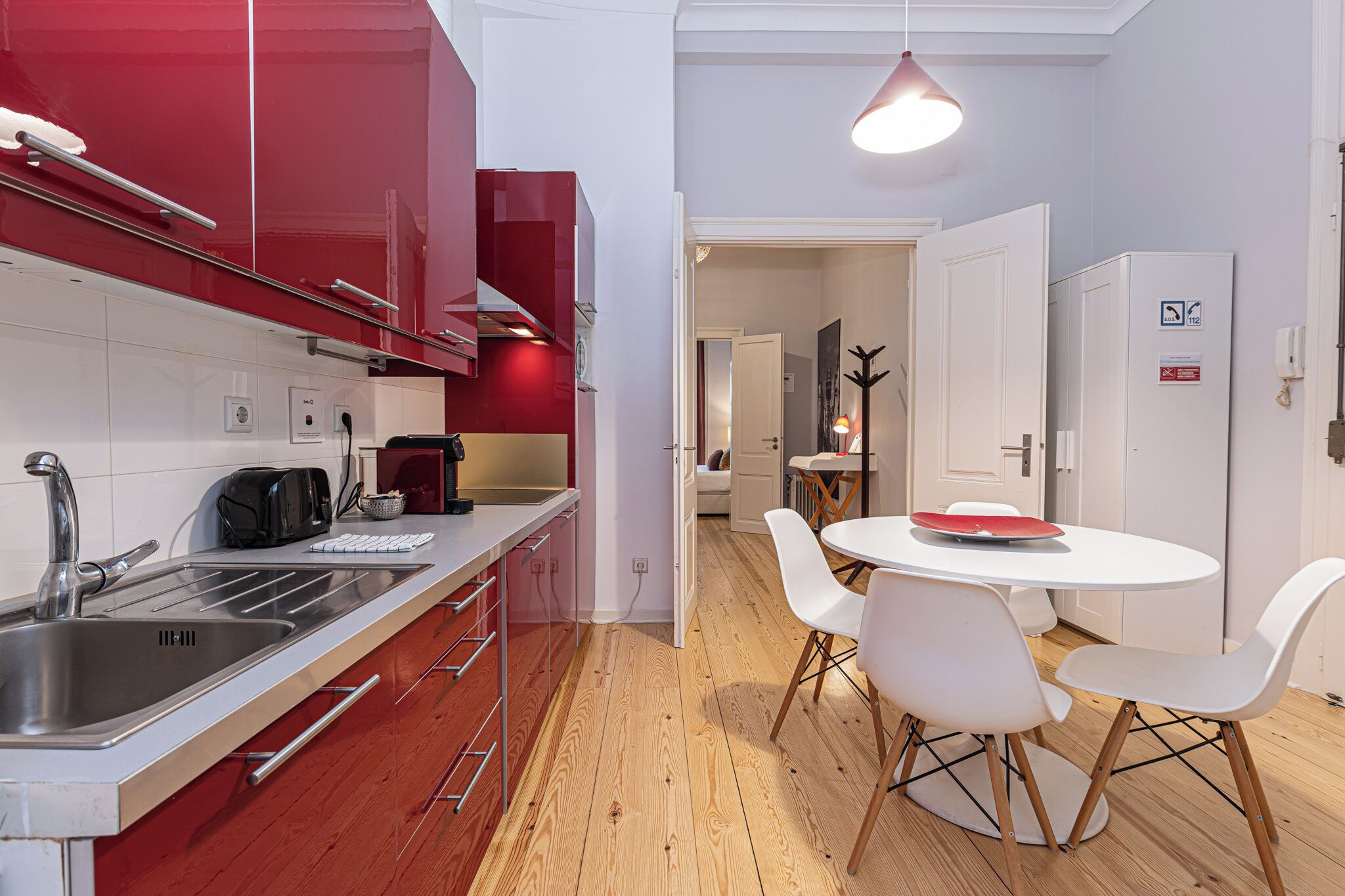 Une cuisine moderne avec un évier en acier inoxydable, des armoires rouges et un plancher en bois. On y trouve également un coin repas avec une table et des chaises blanches.