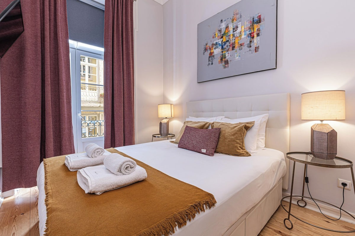 Quarto de hotel com cama, duas luminárias de cabeceira, janela com cortinas e vista para a cidade.