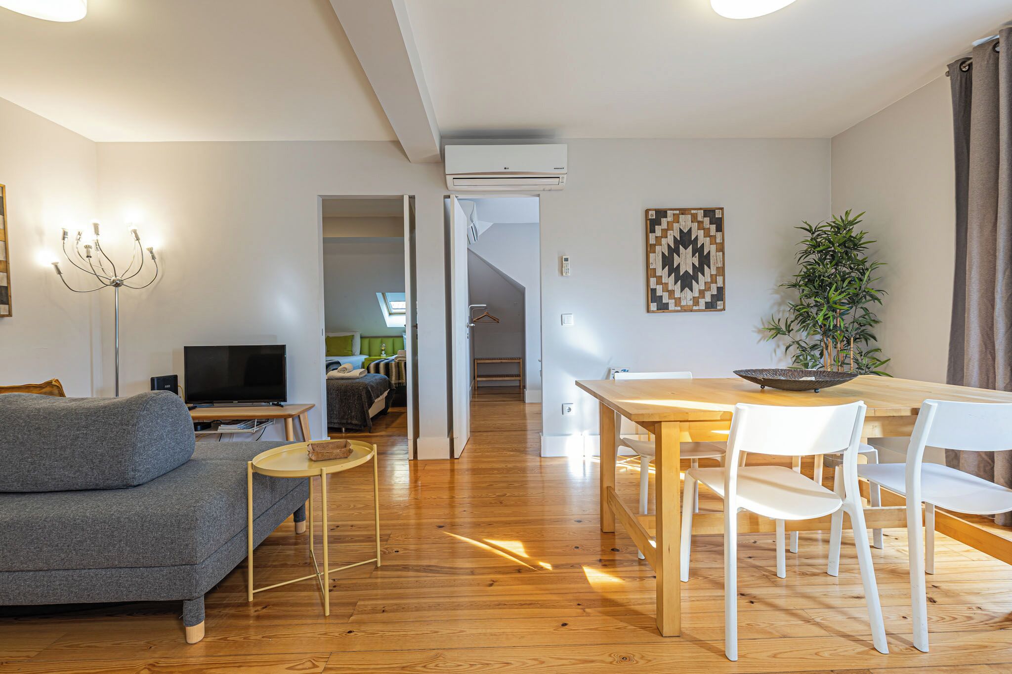 Ein modernes Wohnzimmer mit grauem Sofa, Holzboden, einem Esstisch mit weißen Stühlen und einem Fernseher auf einem Schrank.
