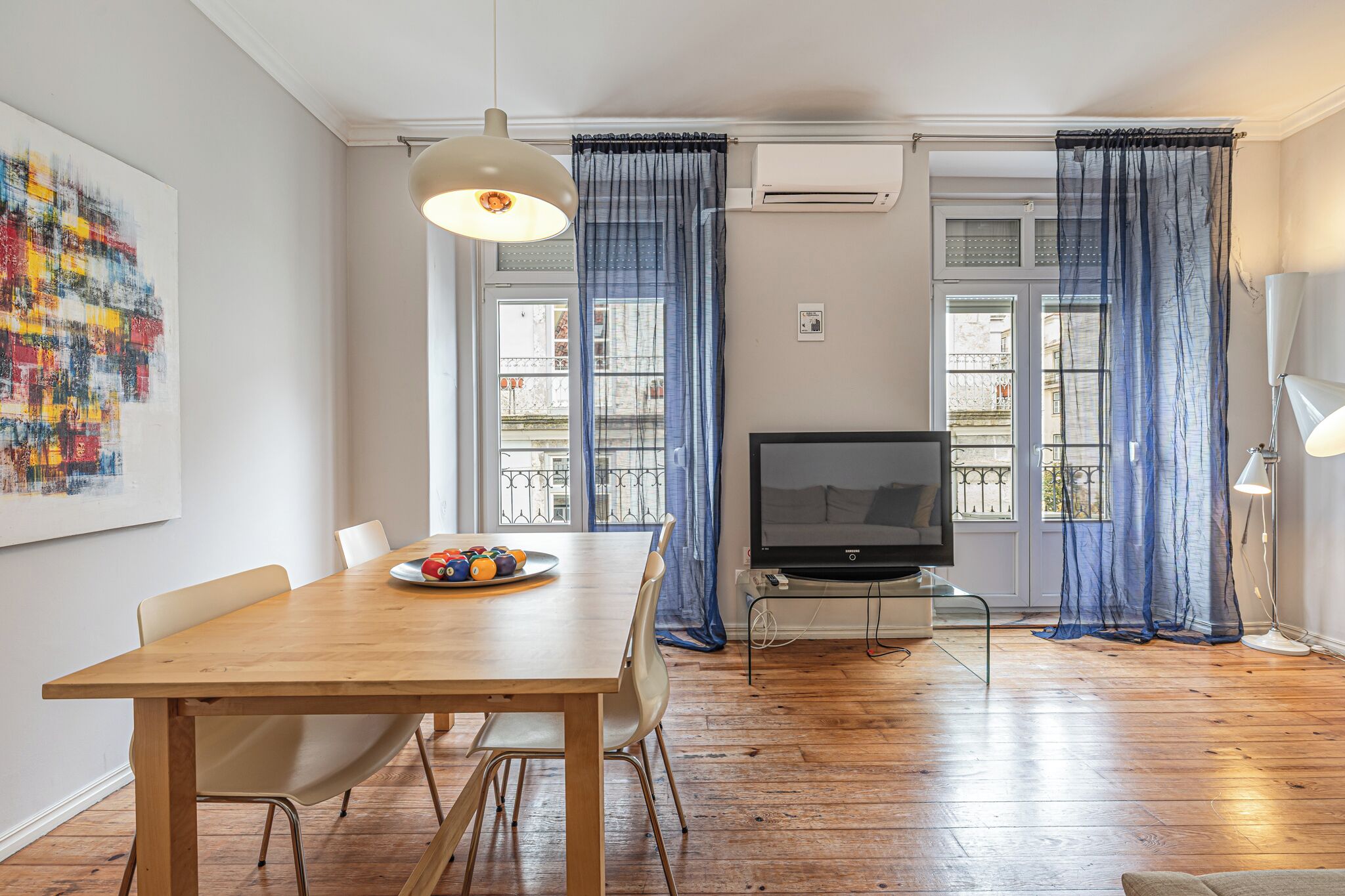 Een moderne woonkamer met een houten eettafel, een flatscreen televisie en een groot raam met blauwe gordijnen.