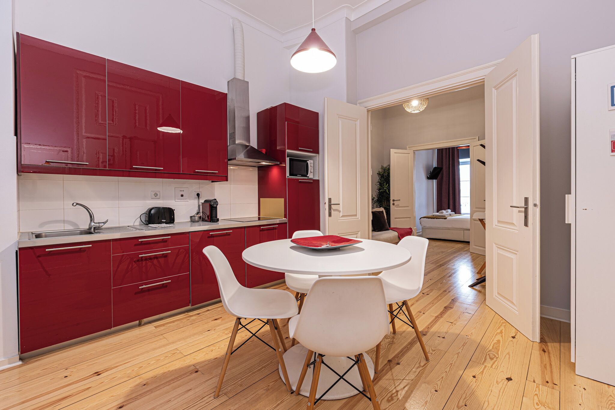 A modern kitchen with red cabinets, a white dining table with four chairs, and a wooden floor.