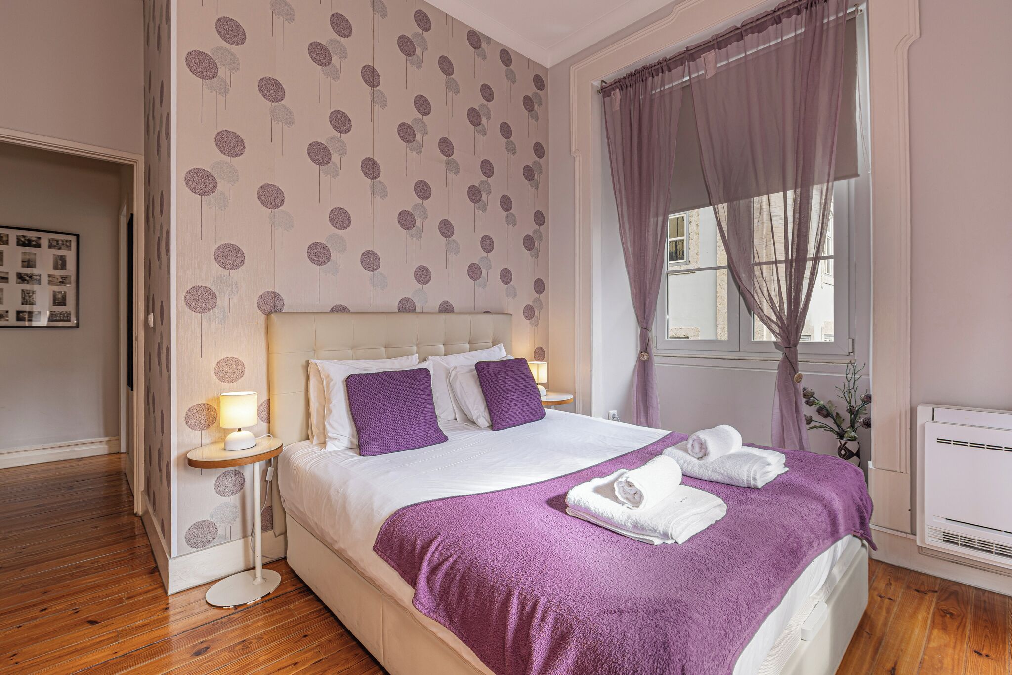 Una cama bien hecha con una manta y almohadas moradas, una lámpara de noche y una ventana con cortinas.