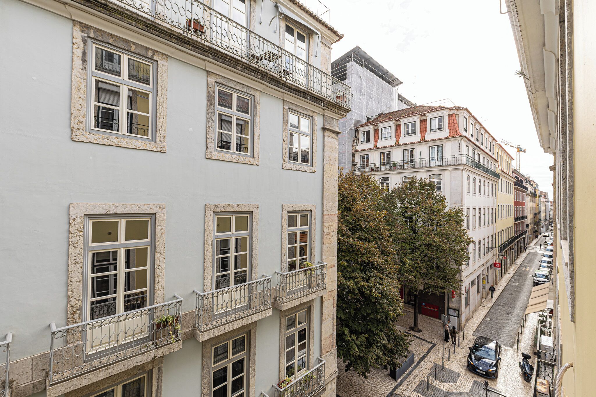 A multi-story building with balconies, a cobblestone street, and parked cars.