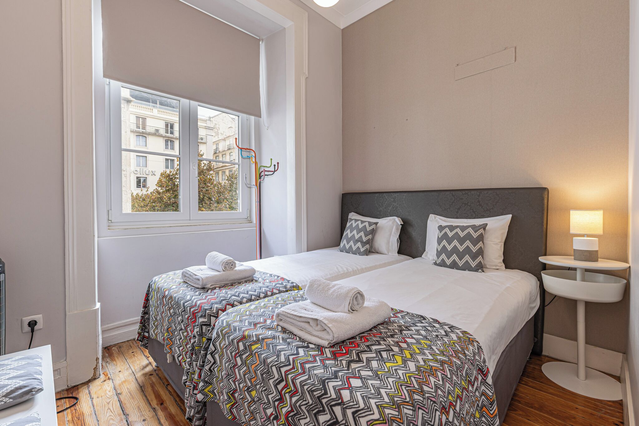 A hotel room with two beds, a window with a view of buildings, a bedside table with a lamp, and a radiator.