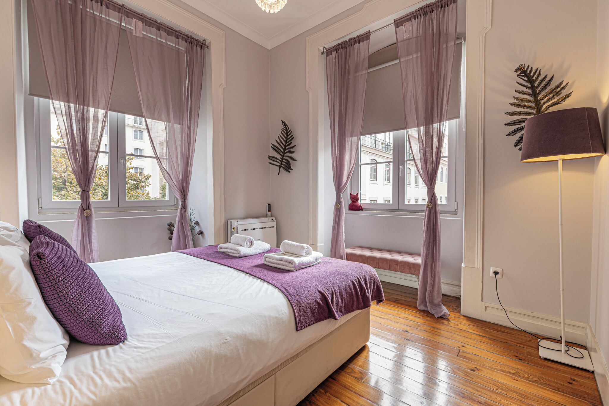 A bedroom with a bed, curtains, a bedside table, a lamp, and a window with a view of buildings.