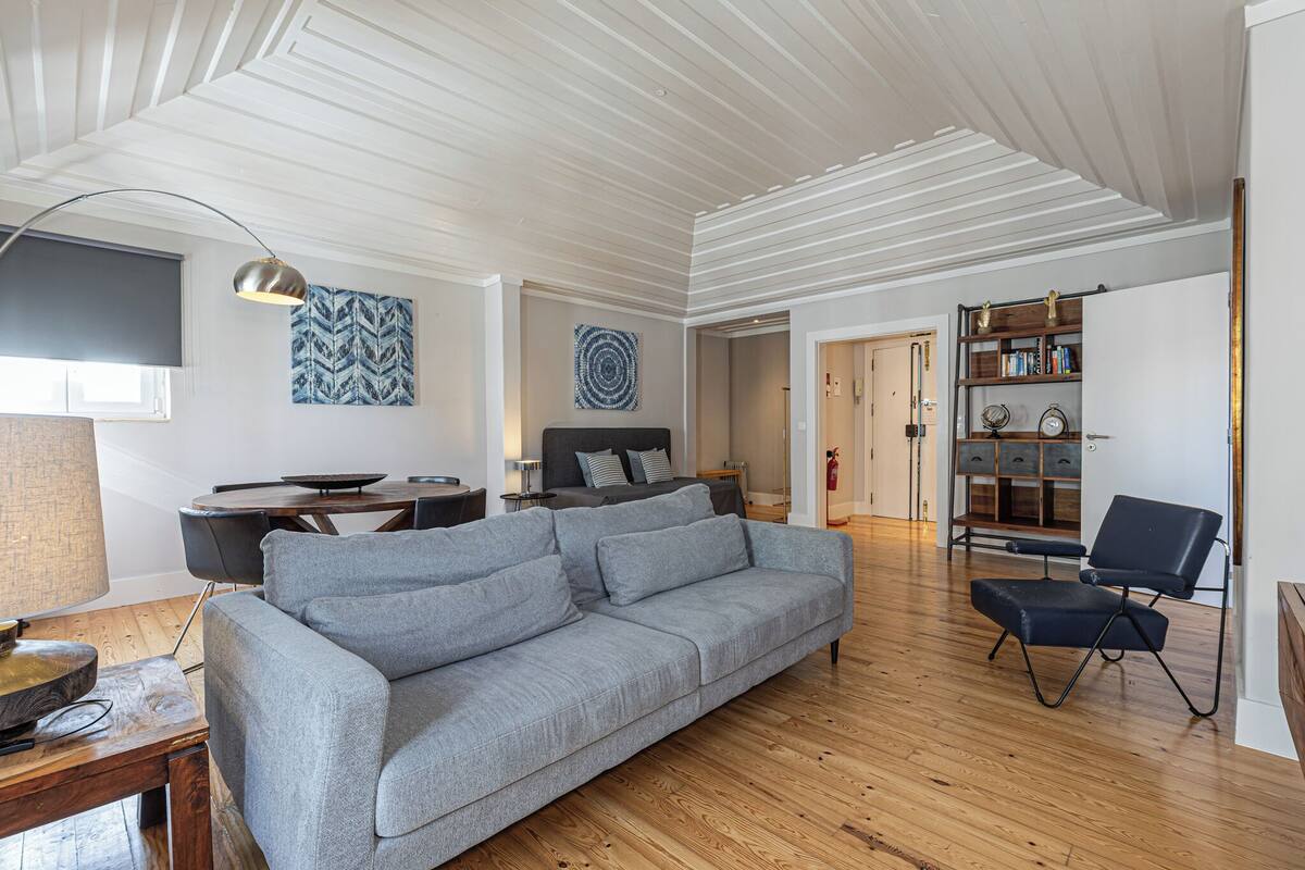 Uma sala de estar moderna com um sofá cinzento, soalho de madeira, uma mesa de jantar e uma estante de livros.