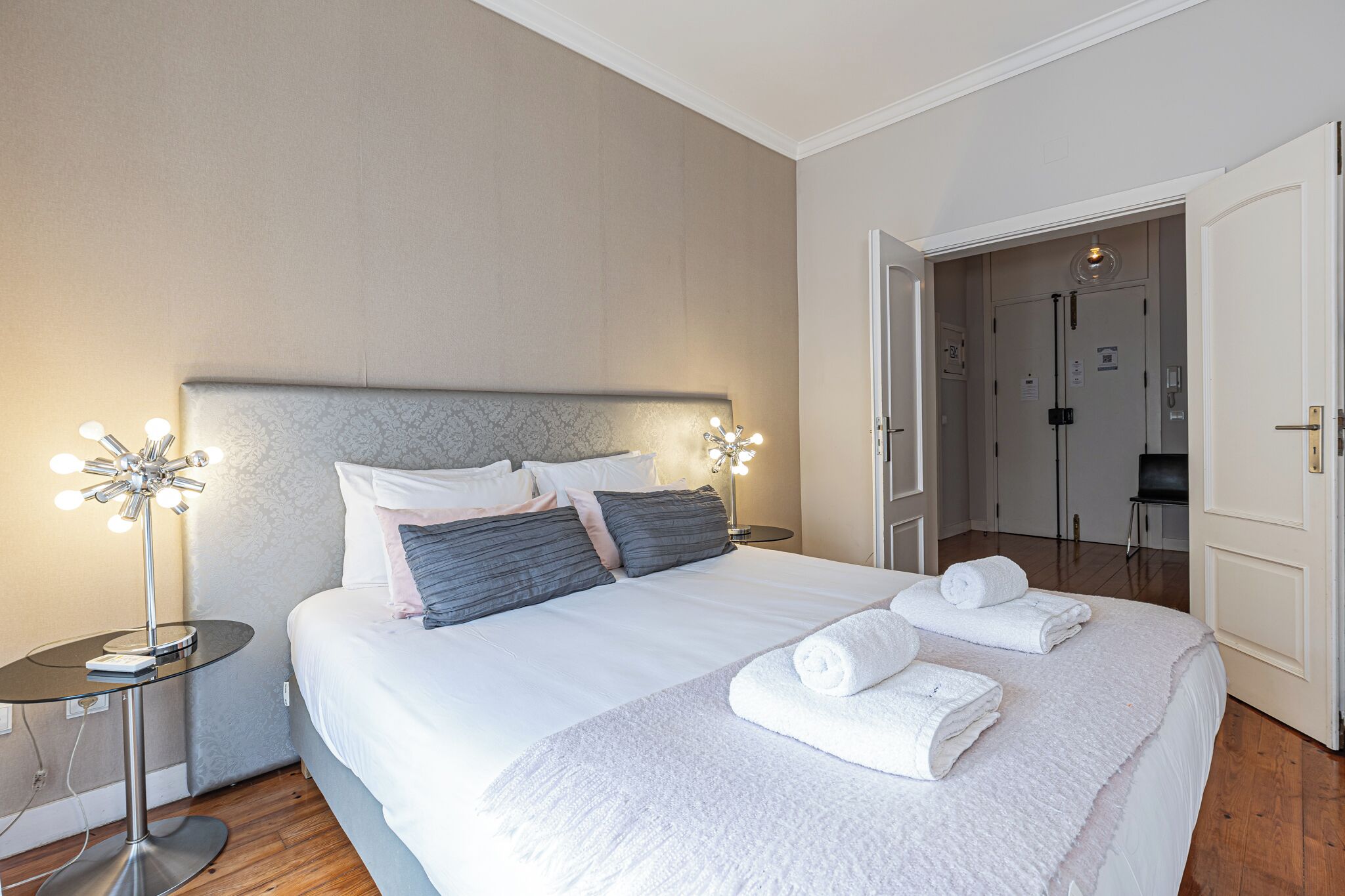 A neatly made bed with white linens and two rolled towels, a bedside lamp, and a door leading to a bathroom.