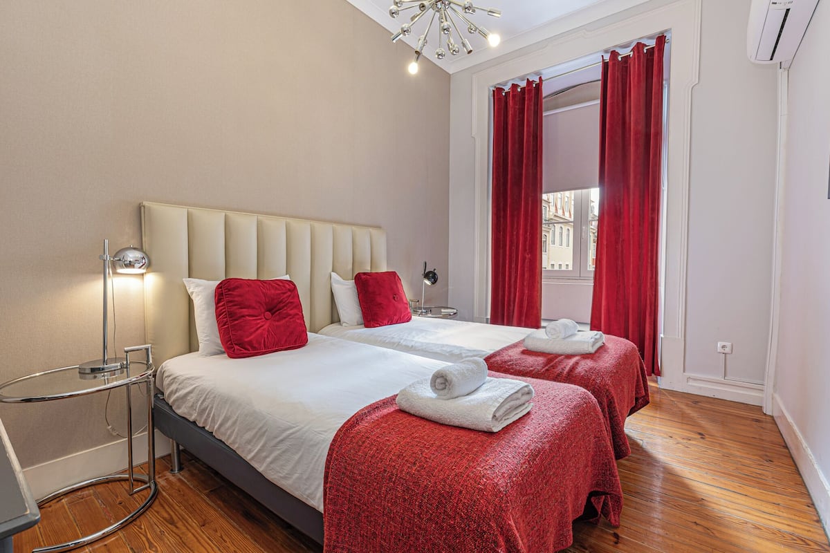 Quarto de hotel com duas camas, roupa de cama vermelha e branca, mesa de cabeceira com abajur e vista para o exterior através de uma janela com cortinas vermelhas.