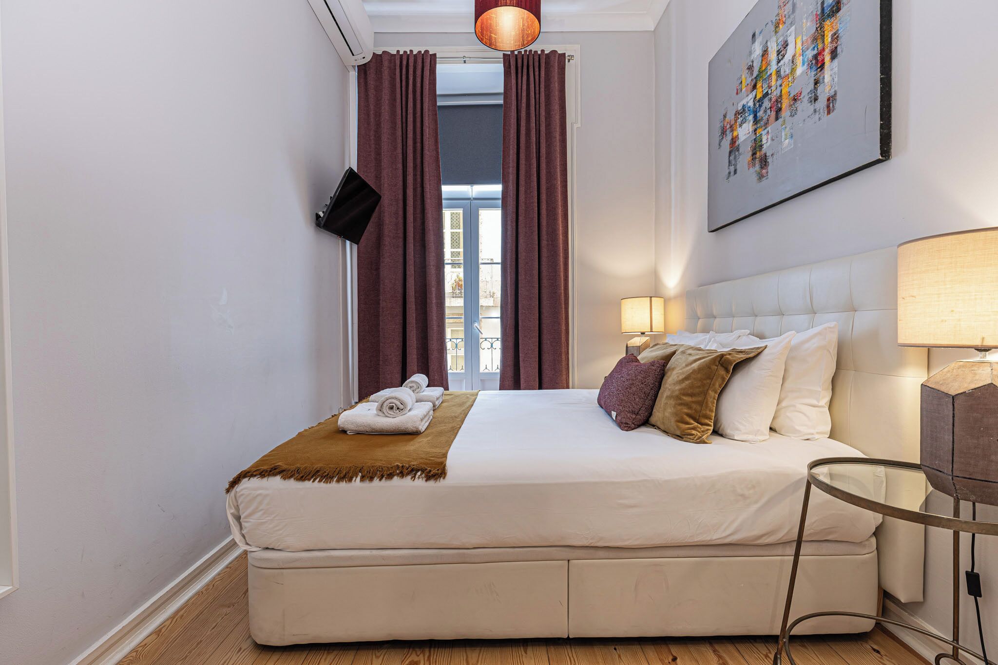 Quarto de hotel com uma cama grande, televisão de tela plana na parede, mesa de cabeceira com abajur e vista para a cidade pela janela.