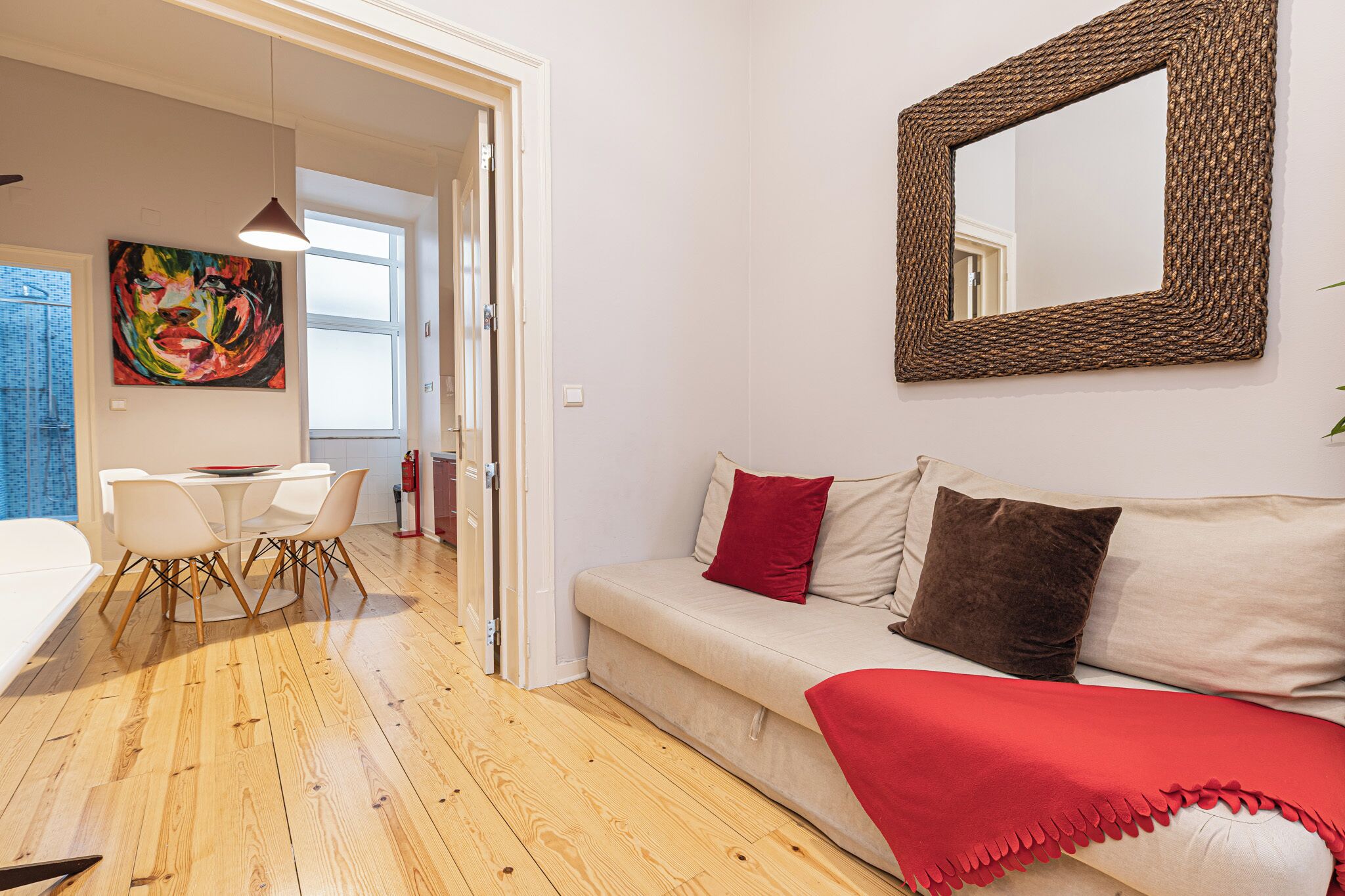 A modern living room with a sofa, red cushions, a red throw blanket, a wooden floor, a dining table with chairs, and a colorful abstract painting on the wall.