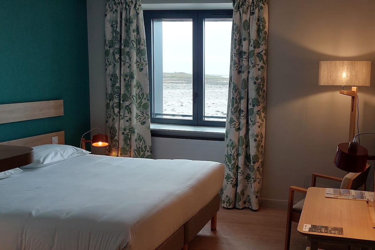 Habitación de hotel con cama, escritorio, lámpara y ventana con cortinas.