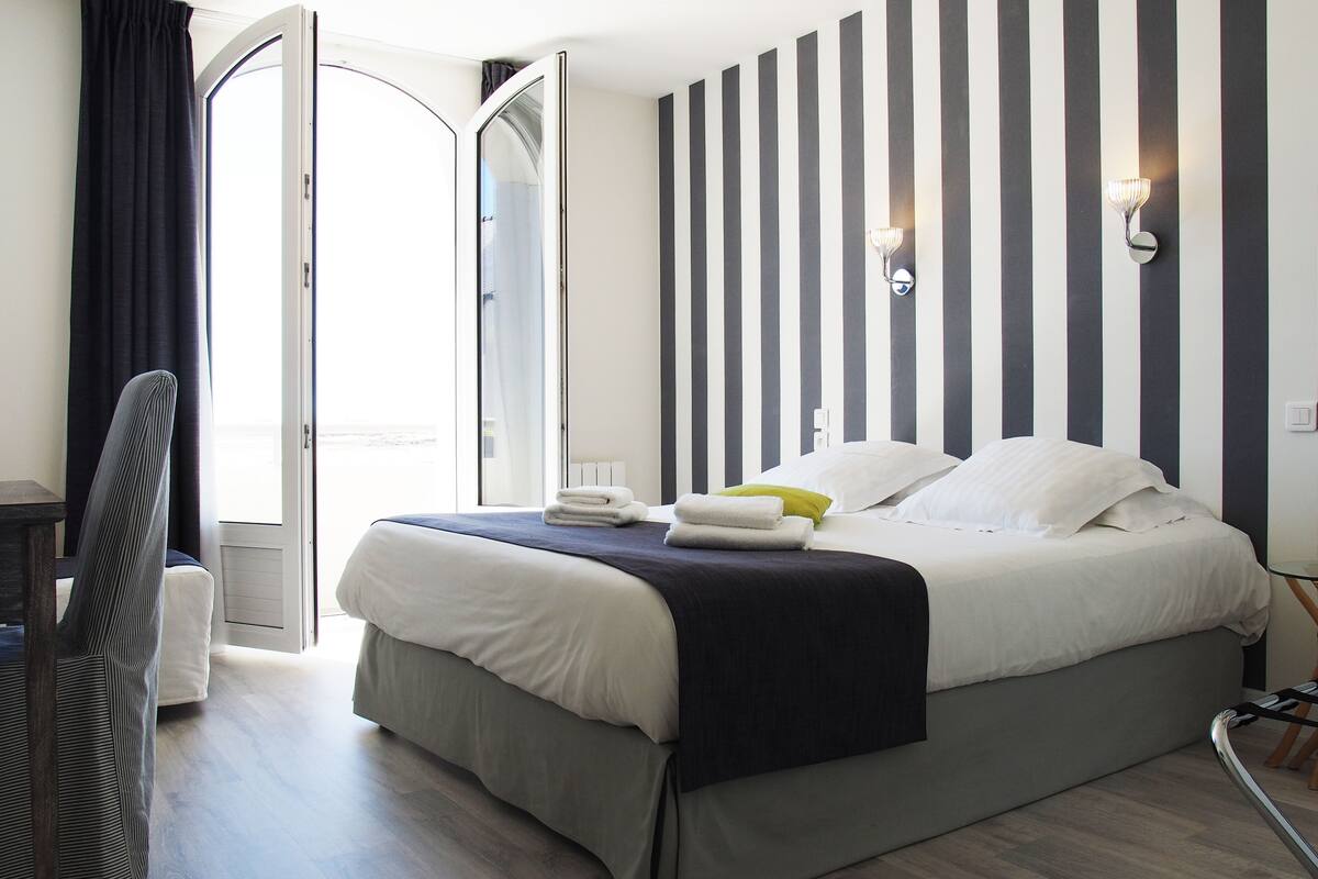 Habitación de hotel con cama, una silla, una mesita de noche, una lámpara de pared y vistas a través de una puerta abierta.