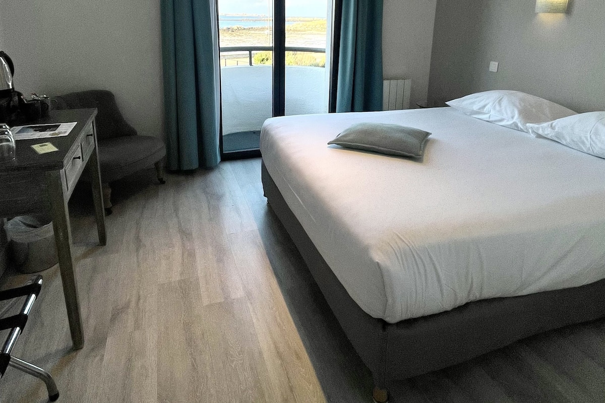 Habitación de hotel con cama, escritorio, silla, ventana con cortinas y vista al exterior.