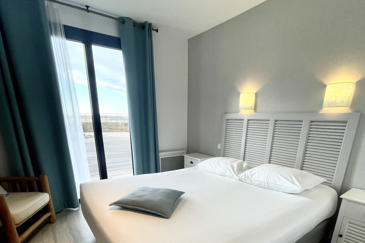 Habitación de hotel con una cama grande, una silla de madera, una mesita de noche y vistas al exterior a través de una puerta corredera.