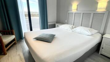 A hotel room with a large bed, a wooden chair, a nightstand, and a view of the outdoors through a sliding door.