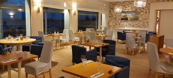 Restaurante com mesas redondas, cadeiras azuis e brancas, e parede de pedra com uma pintura.