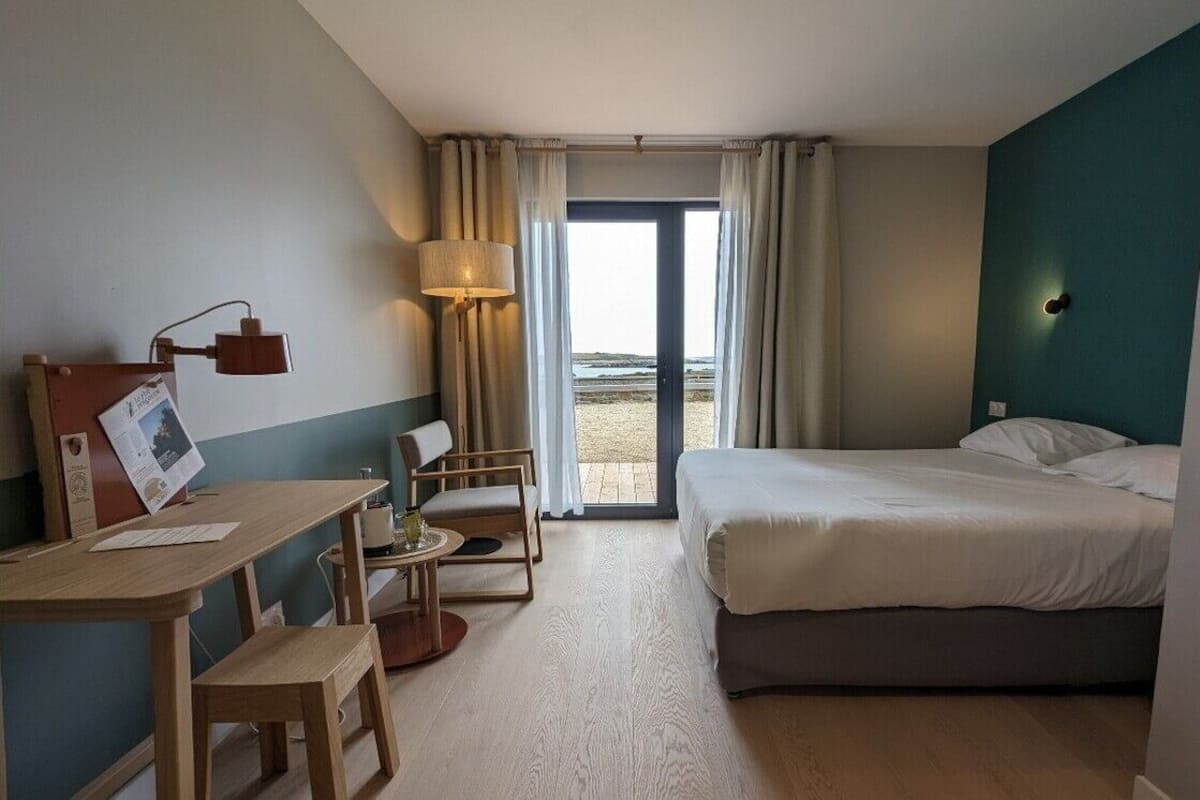 Habitación de hotel con cama, escritorio, silla, lámpara y vista a la playa a través de un gran ventanal.
