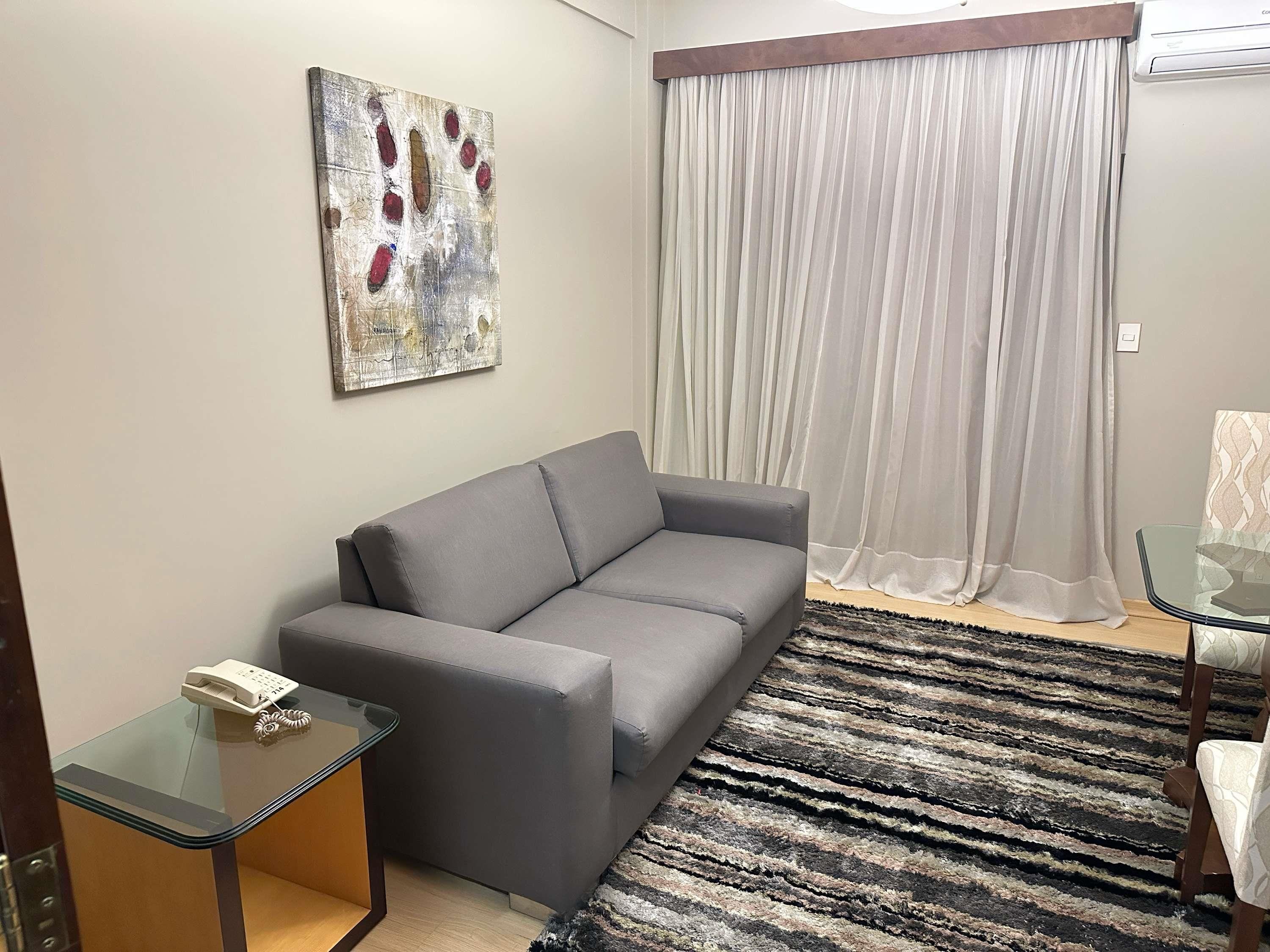 A living room with a grey sofa, a glass coffee table with a telephone, a patterned rug, and a large window with sheer curtains.