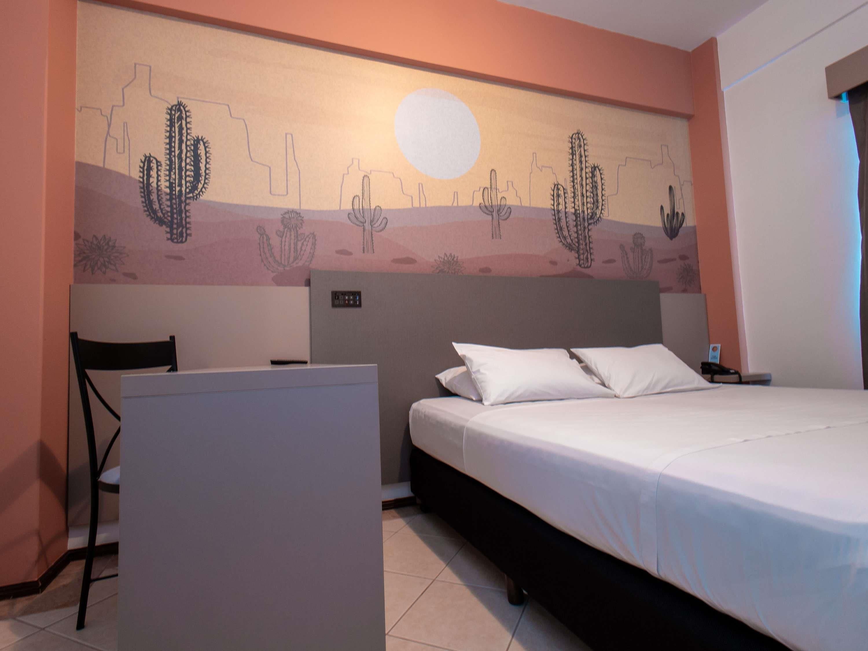 Een hotelkamer met een bed, een bureau en een muurschildering van cactussen en een woestijnlandschap.