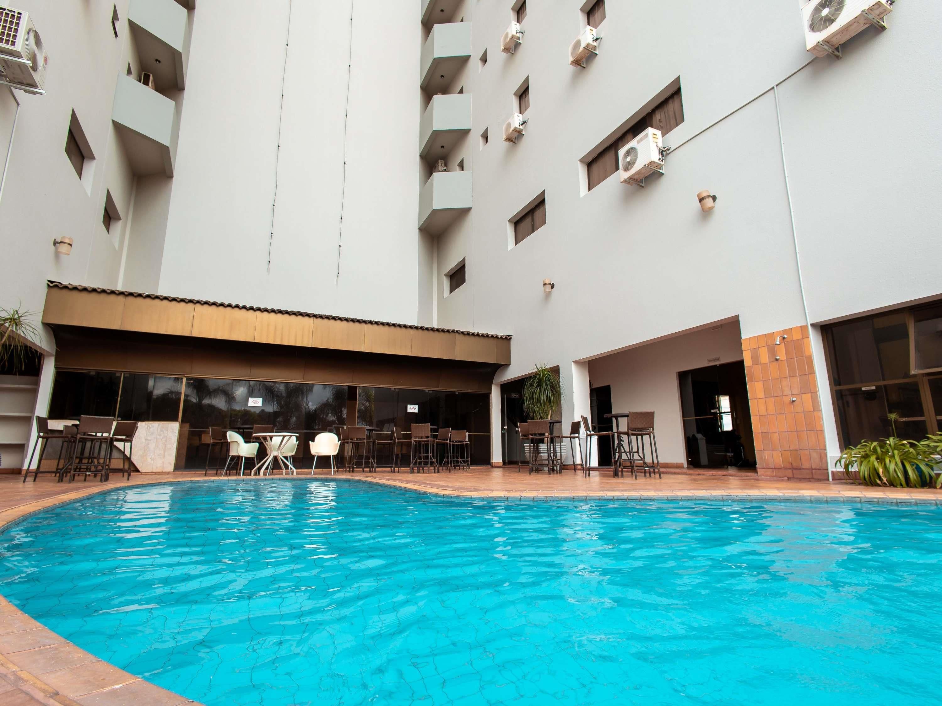 A swimming pool with lounge chairs and tables, surrounded by a multi-story building with balconies and air conditioning units.