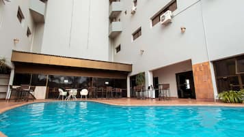 Una piscina con sillas de descanso y mesas, rodeada por un edificio de varios pisos con balcones y unidades de aire acondicionado.