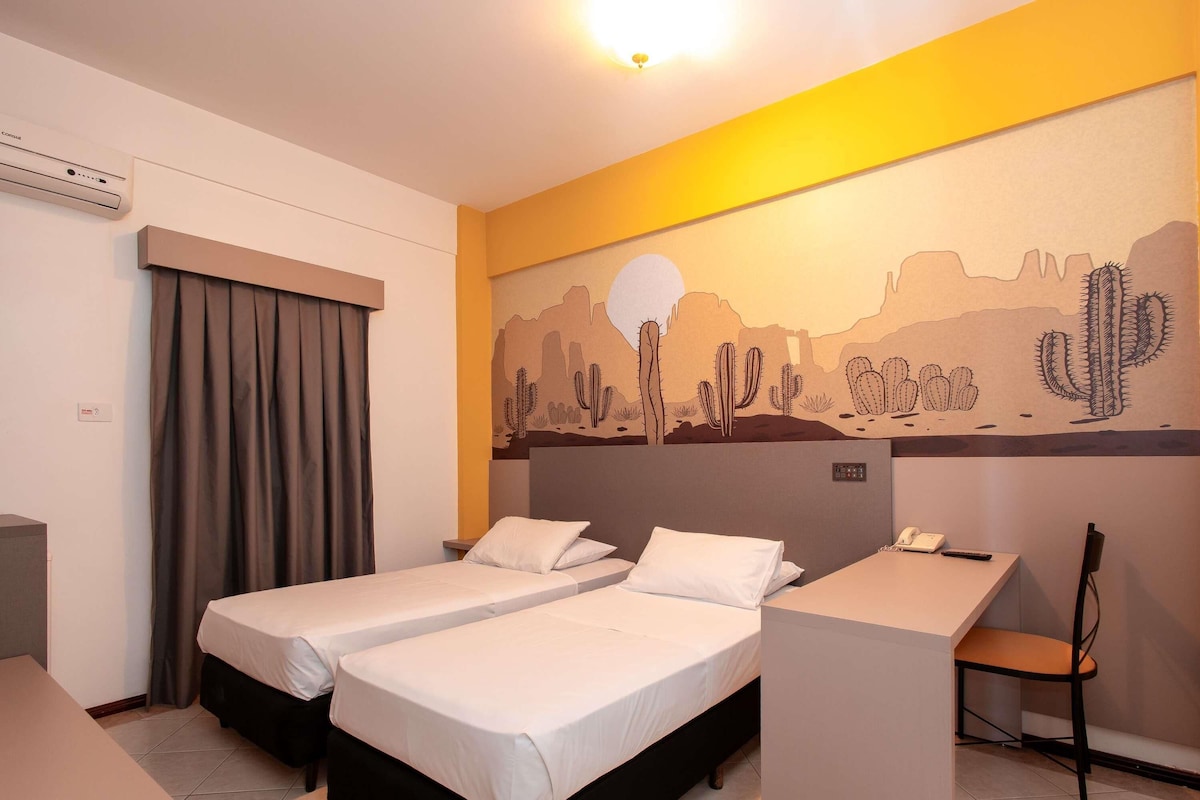 A hotel room with two beds, a desk, a chair, and a desert-themed mural on the wall.