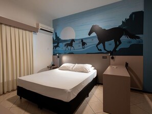 A hotel room with a bed, a nightstand, a desk, an air conditioning unit, and a wall mural of horses.