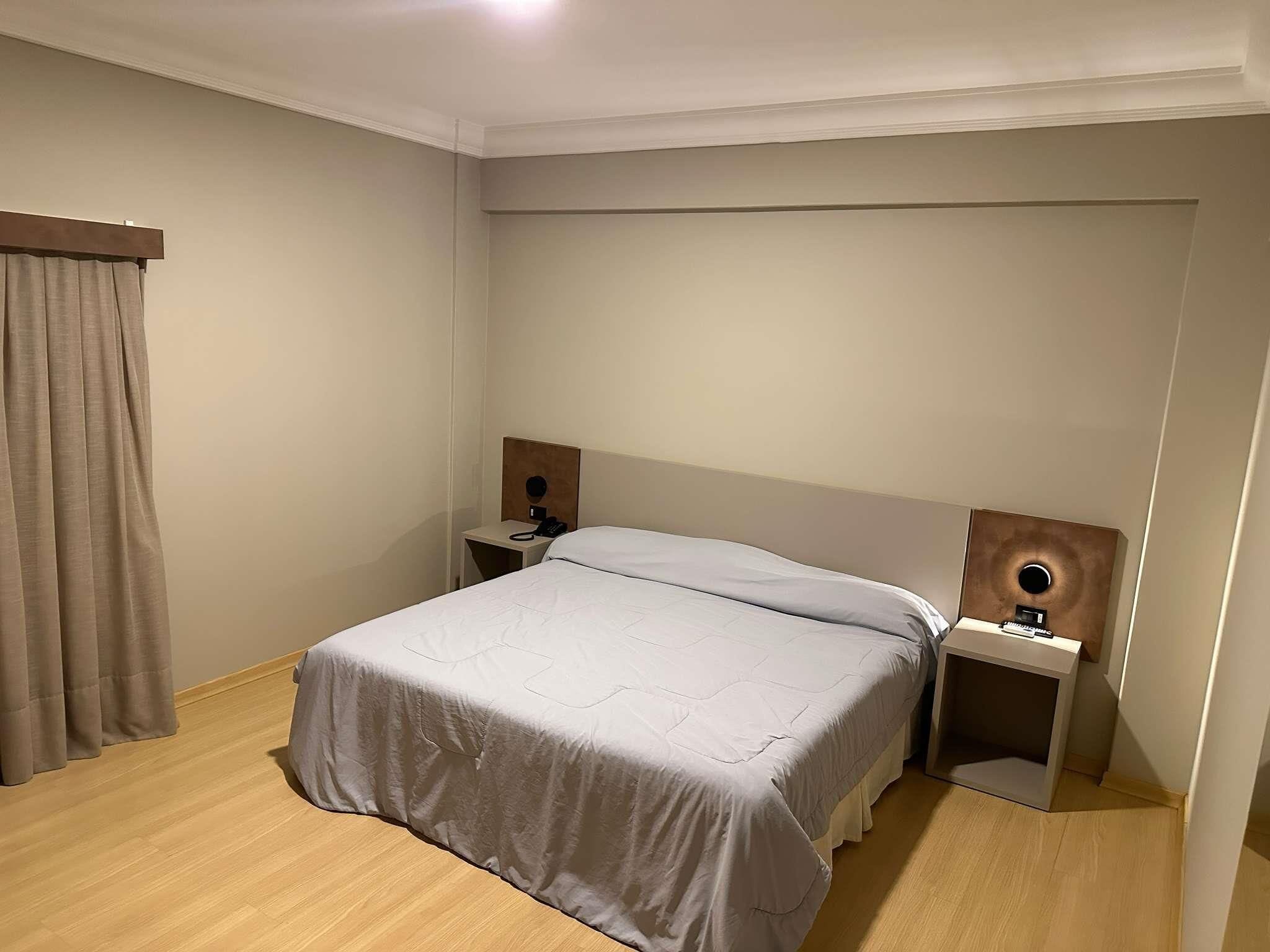 Hotellihuone, jossa on sänky, yöpöytä, jossa on puhelin, seinään kiinnitetty valaisin ja verho.