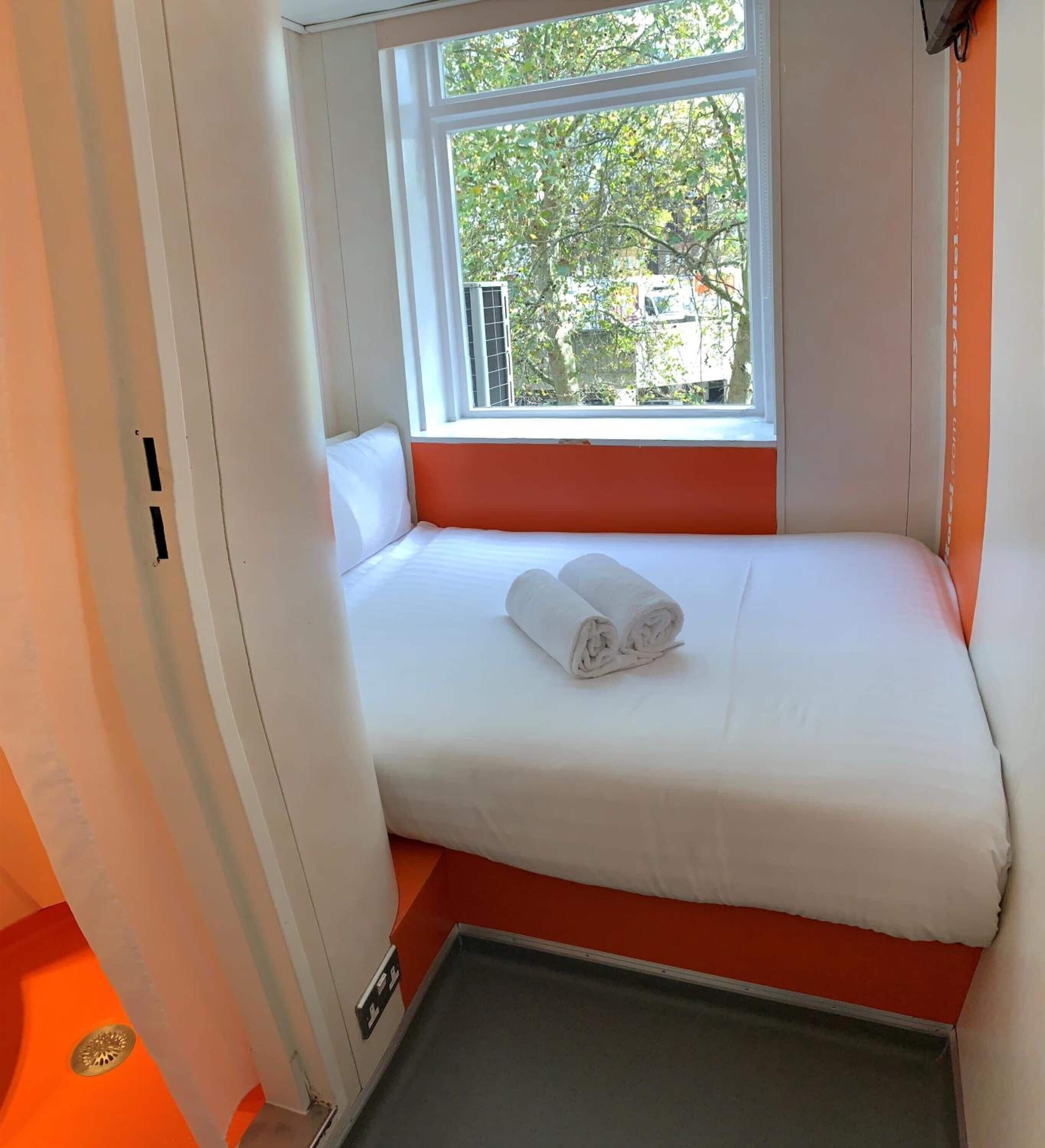 Ein kleines Zimmer mit einem Einzelbett, Fenster, weißer Bettwäsche und einem aufgerollten Handtuch auf dem Bett.