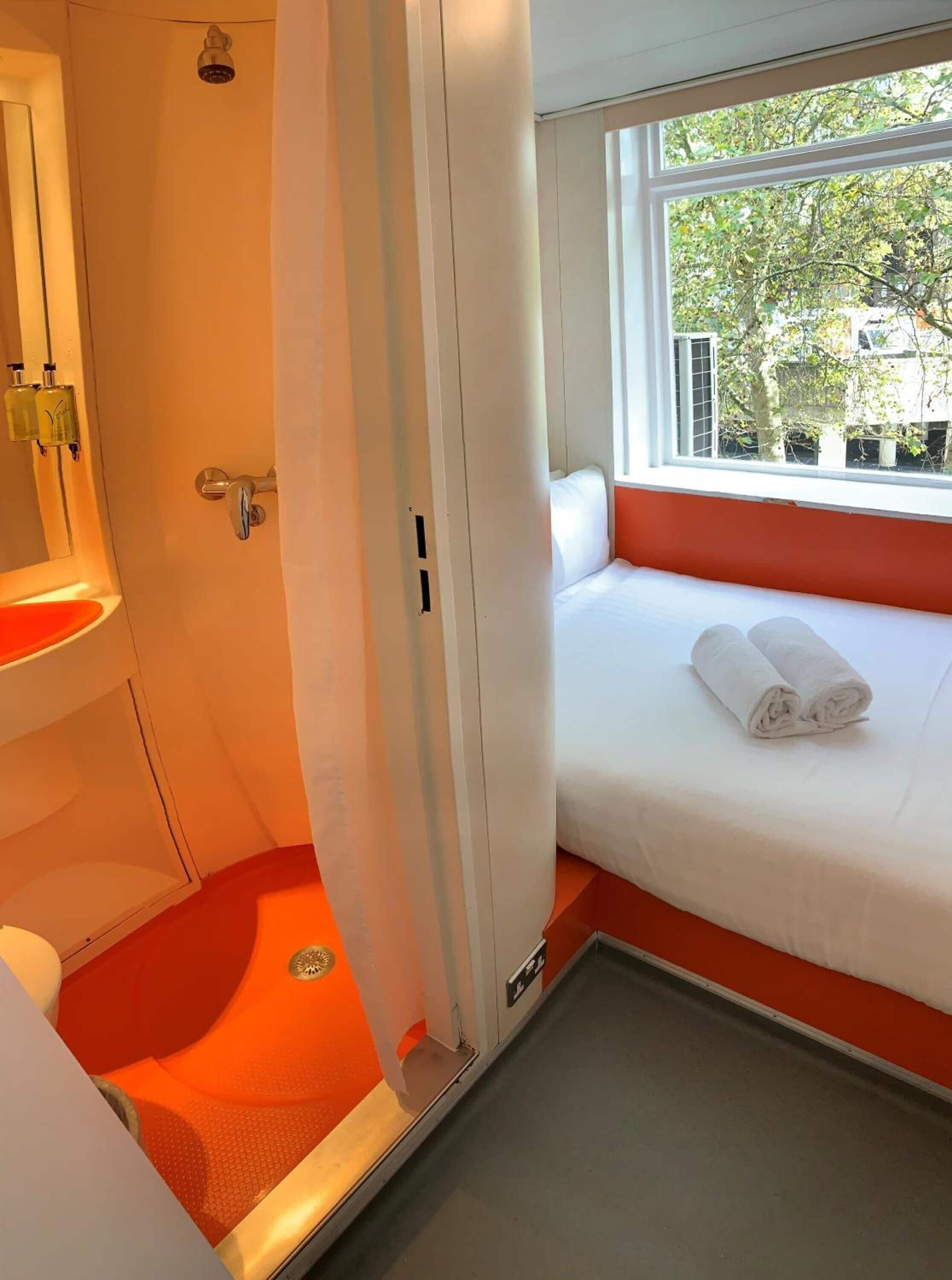 A hotel room with a bed, a window with a view of trees, a bathroom with a sink and mirror, and a shower area.