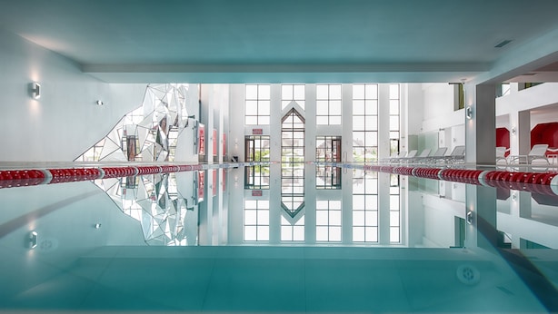 A modern indoor swimming pool with clear glass walls, a geometric ceiling design, and a view of a building outside. - Wish Aqua & Spa Resort (Vyshen'ky)