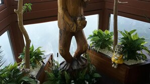 Una escultura de madera de una figura en un entorno boscoso, rodeada de plantas en macetas y una vista de una masa de agua a través de un ventanal grande.