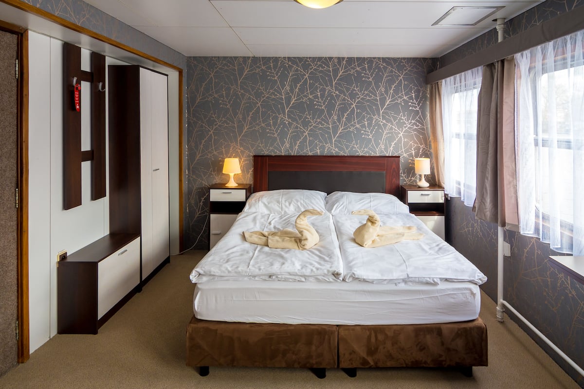 Habitación de hotel con una cama grande, dos mesitas de noche con lámparas, un armario y vistas al exterior a través de cortinas traslúcidas.