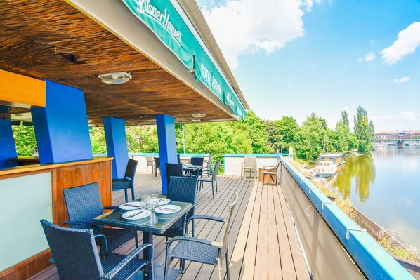 Une terrasse extérieure avec des tables et des chaises, donnant sur une rivière où l’on peut voir des bateaux et un pont en arrière-plan.