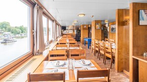 Un vagón comedor con mesas y sillas de madera, ventanas amplias con vistas a un río y la imagen de barcos.