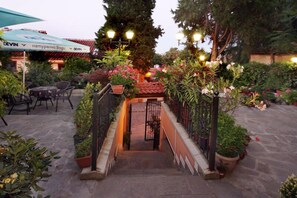 Zona exterior amb terrassa coberta, plantes en testos i escala que condueix a l'entrada d'un edifici.