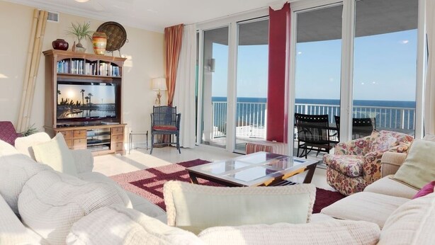 A spacious living room with a large flat-screen TV, a bookshelf, and a comfortable seating area with a view of the sea through large sliding glass doors. - Mustique 901 (Gulf Shores)