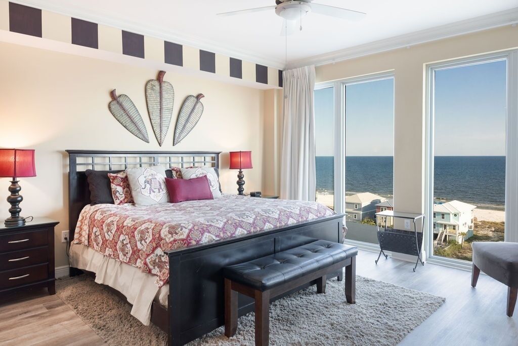 Una camera da letto con un letto grande, due abat-jour, una panca, vista sull'oceano e una ventola a soffitto.