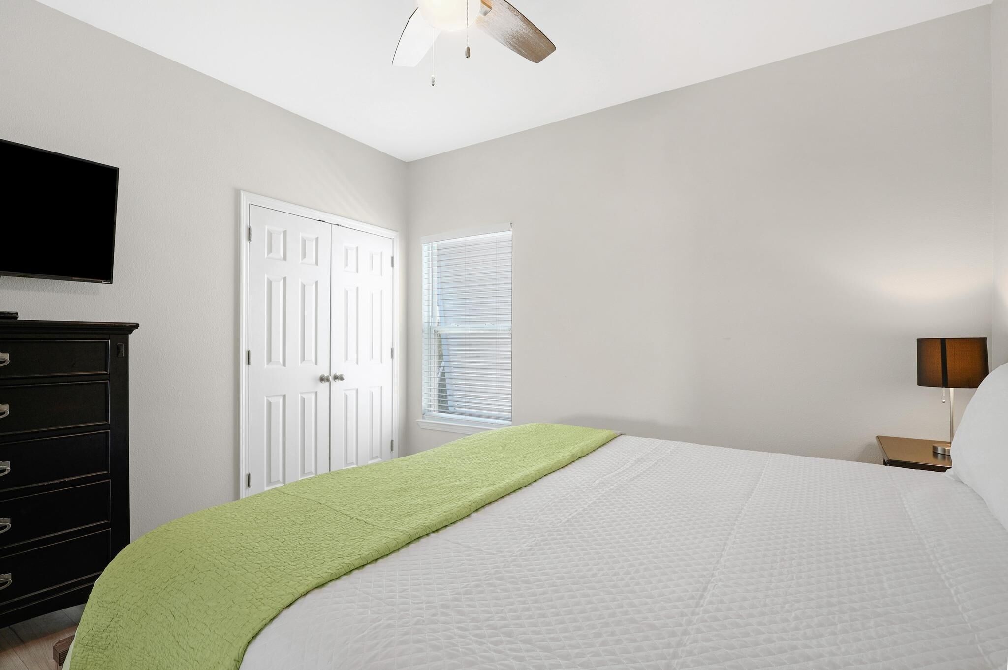 Un dormitorio con cama, cómoda, televisor, puerta y ventana con persianas.