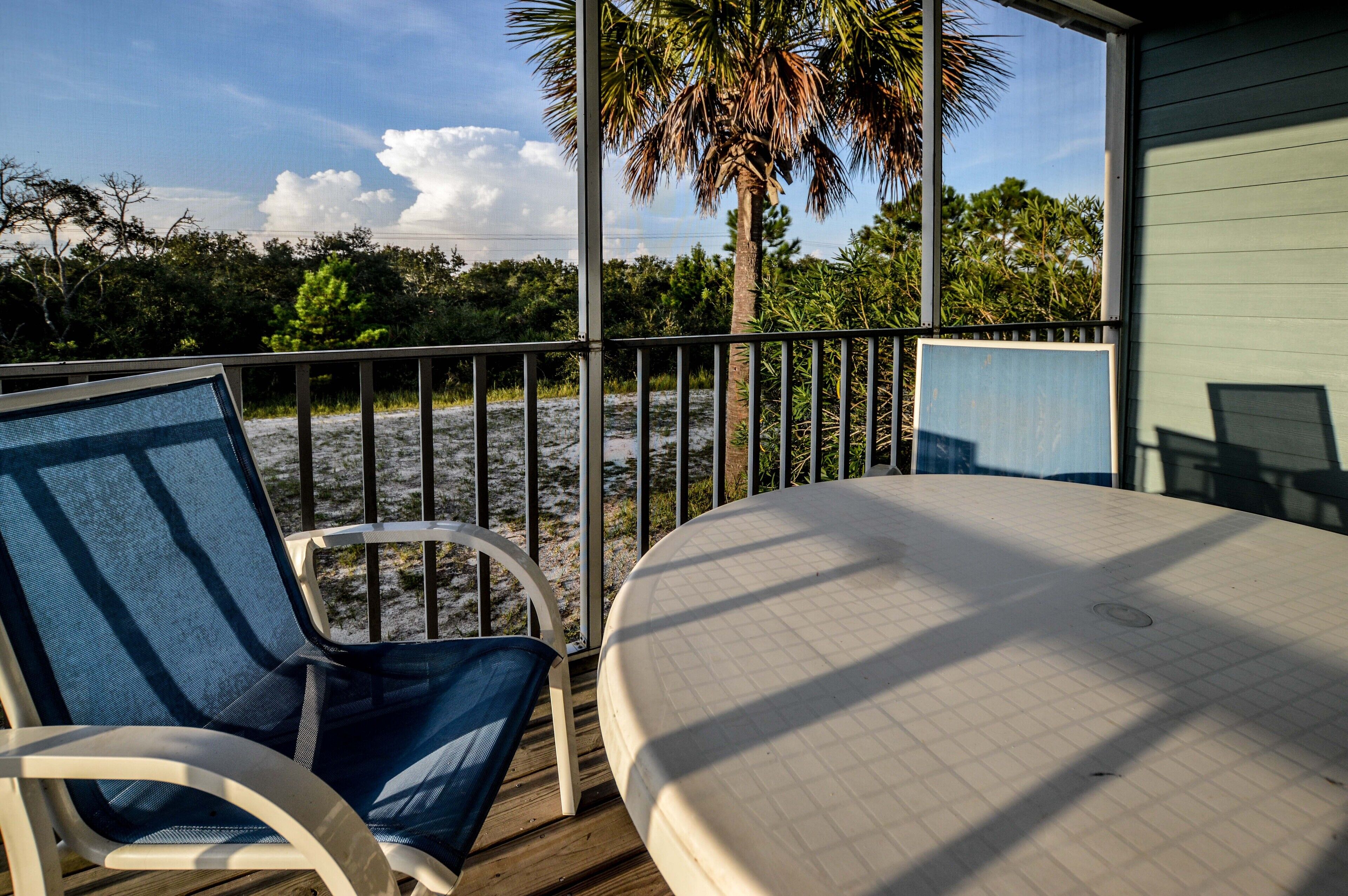 A deck with two chairs, a table, and a view of the beach and palm trees.