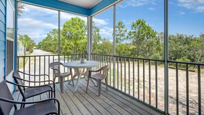 A covered outdoor patio with a table and chairs, large windows, and a view of trees and a path. - The Rookery III Unit 4001 (Gulf Shores)