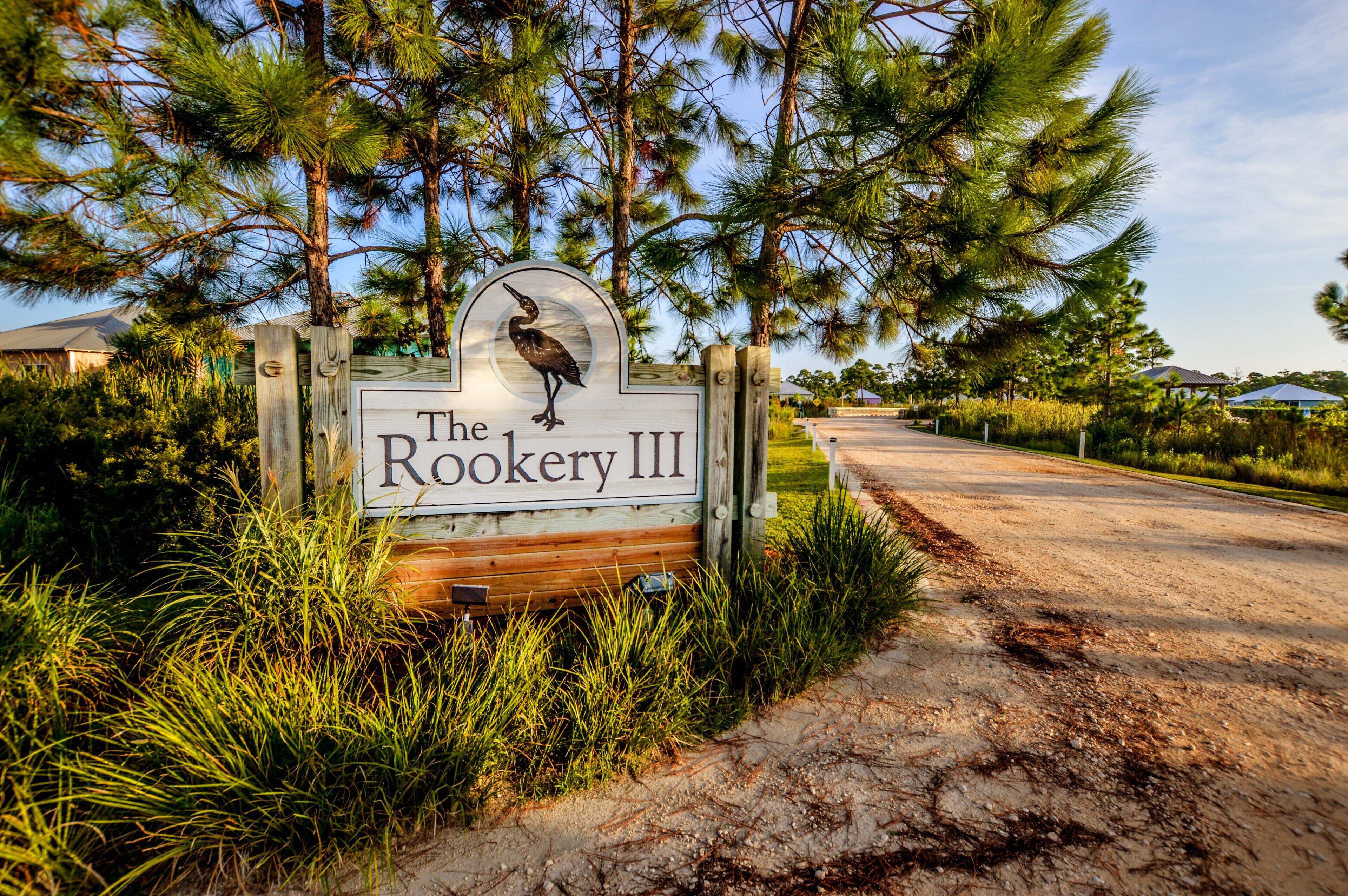 A wooden sign with a bird and the text "The Rookery III" in a natural setting with trees and a dirt path.