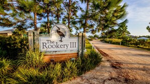 A wooden sign with a bird and the text "The Rookery III" in a natural setting with trees and a dirt path. - The Rookery III Unit 4001 (Gulf Shores)