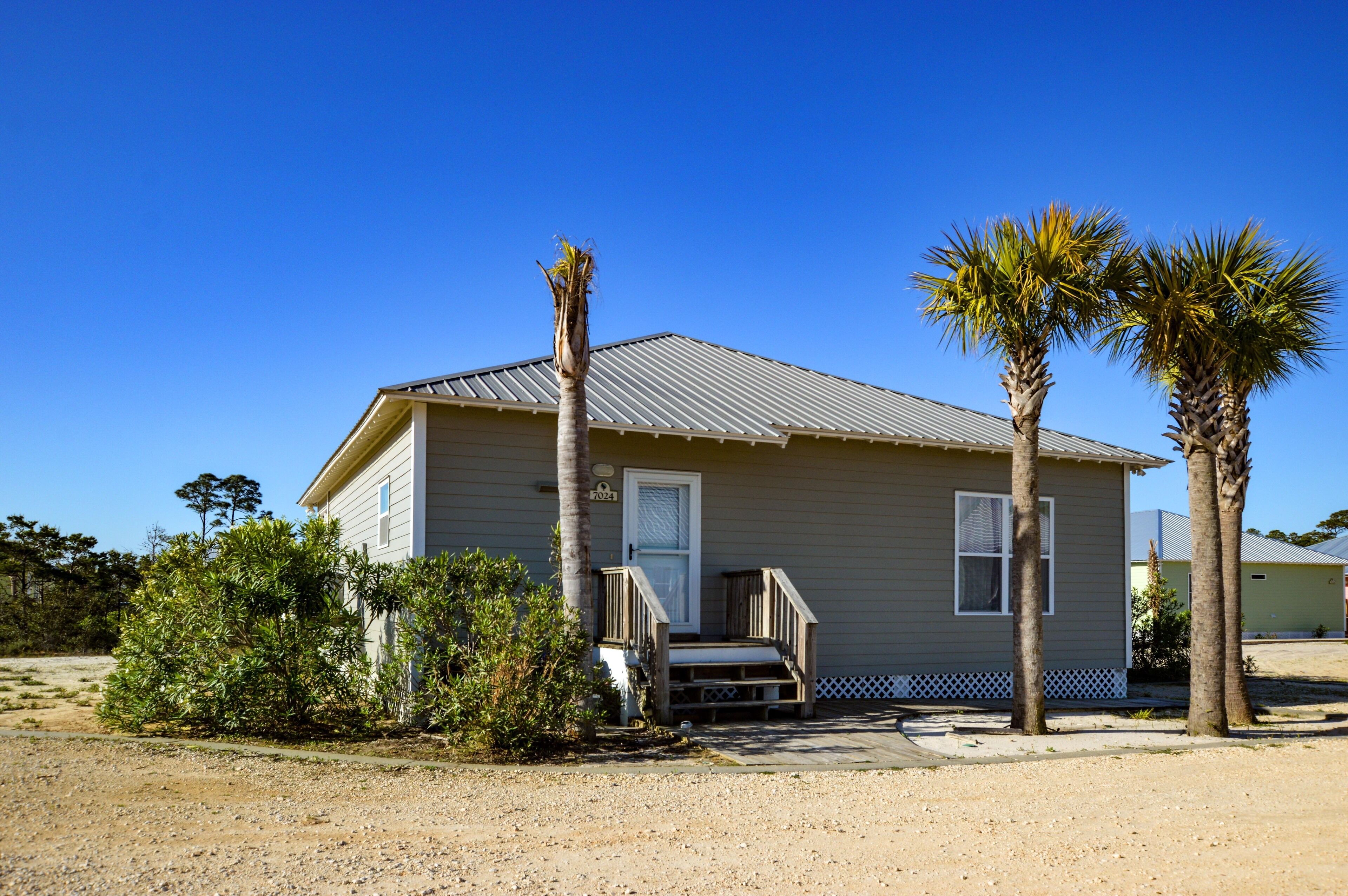 A single-story house with a metal roof, a front porch with a wooden railing, and two palm trees.