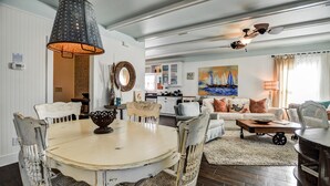 A dining area with a round table and chairs, a kitchen in the background, and a painting of sailboats on the wall.