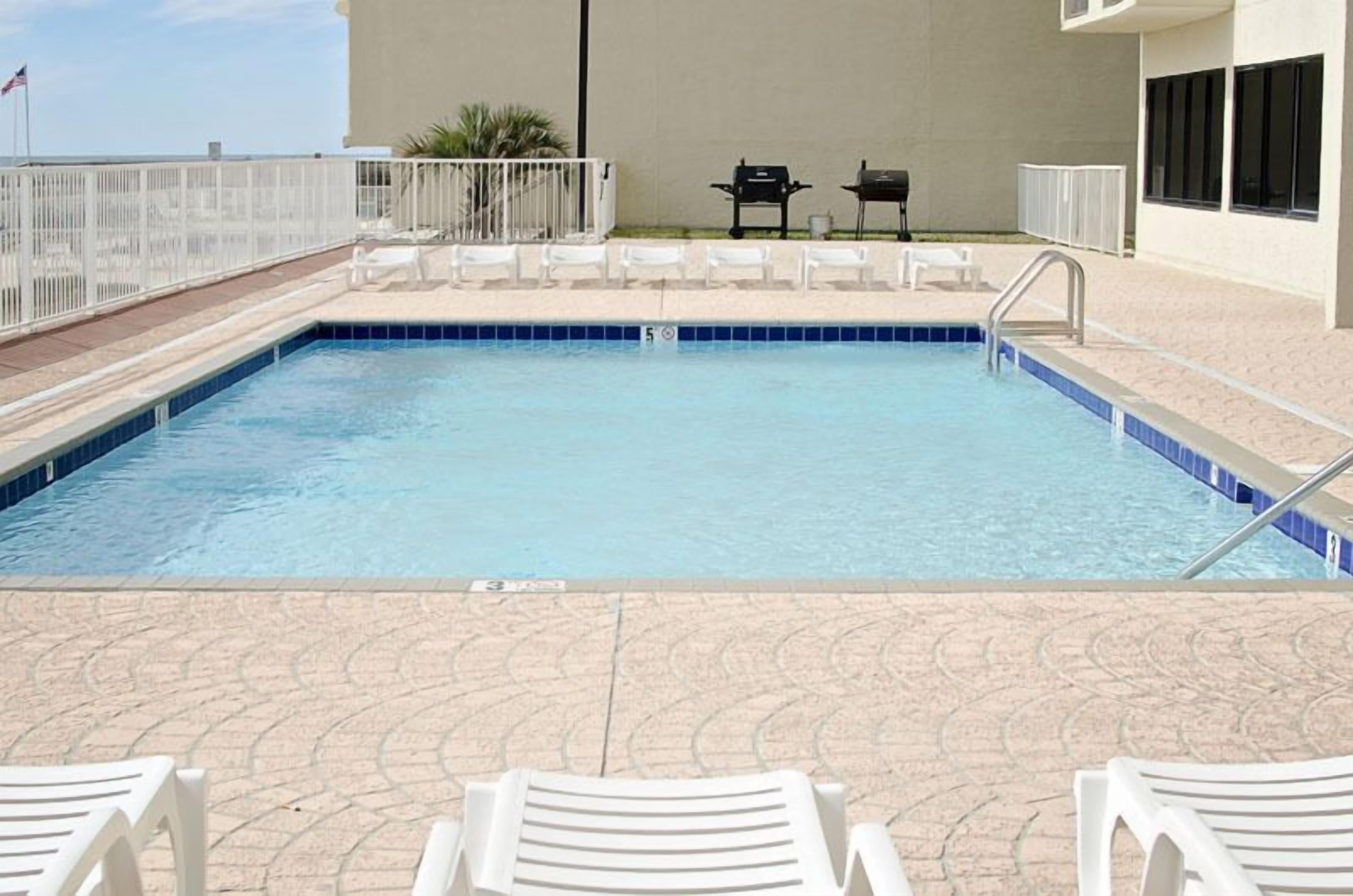 A rectangular pool with a blue tiled edge, surrounded by a white fence, and white lounge chairs on the deck.