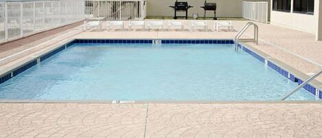 A rectangular pool with a blue tiled edge, surrounded by a white fence, and white lounge chairs on the deck.